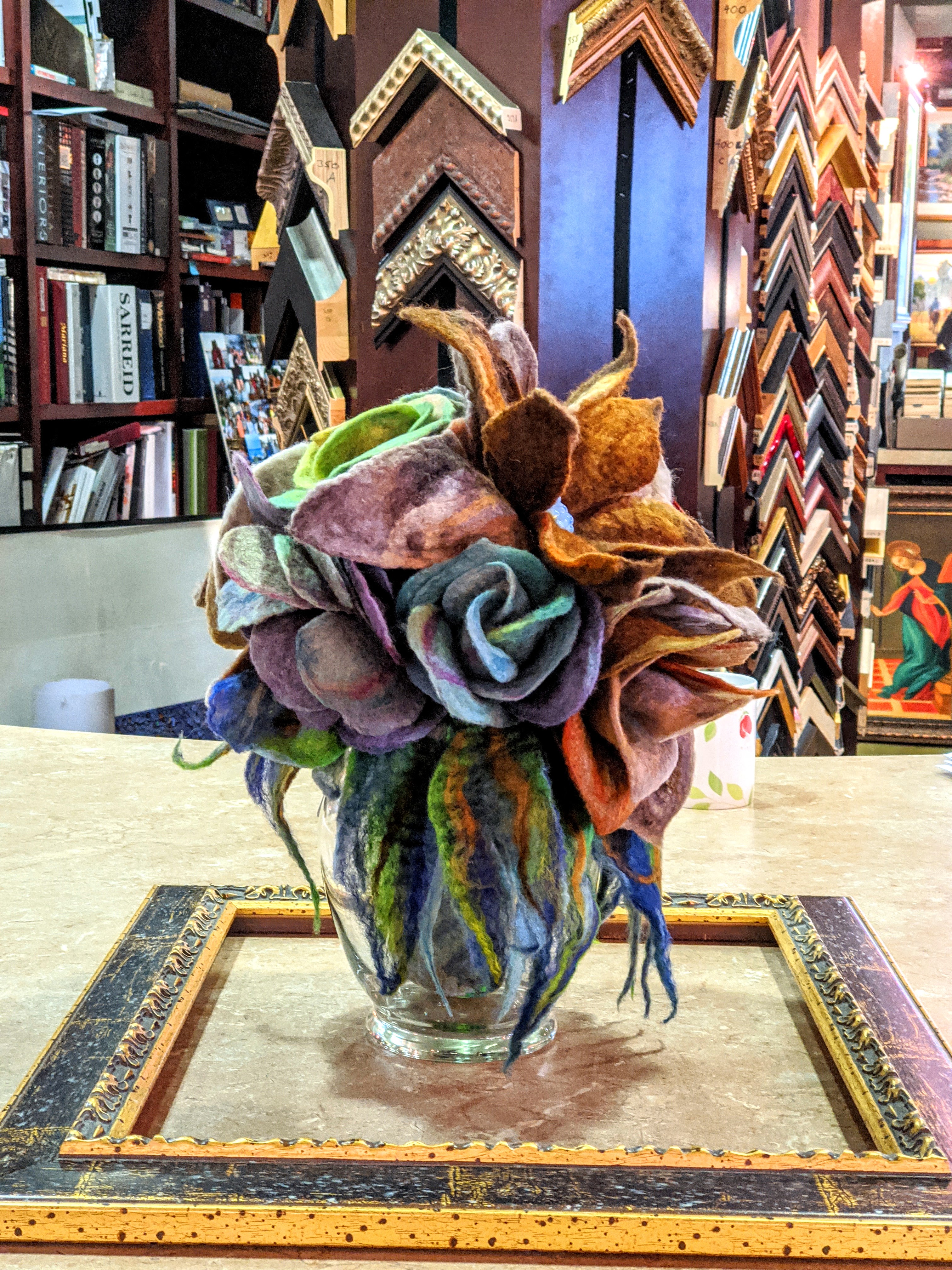 Handcrafted felted wool floral arrangement in a glass vase featuring textured purple, ochre, and blue petals on a framing counter